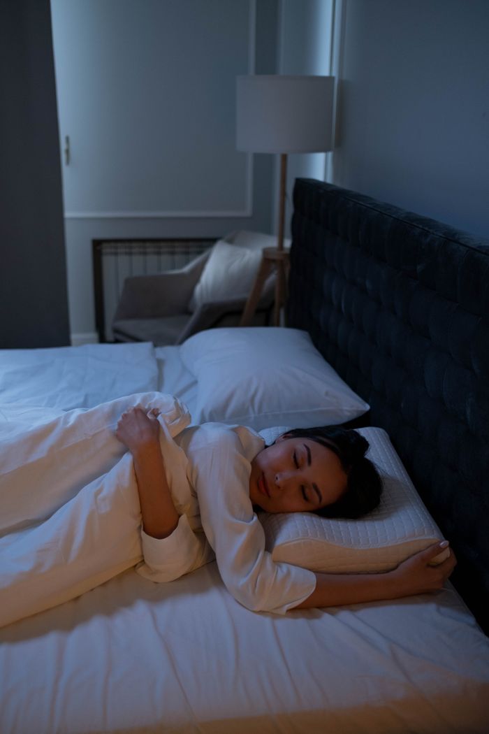 Asian woman peacefully sleeping in a cozy, dimly lit bedroom on white bedding.